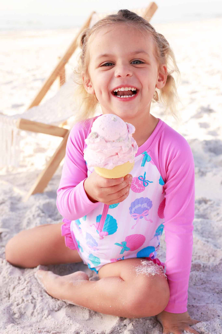 girl sitting on beach eating ice cream wearing a colorful seashell long sleeve rash guard swimsuit 