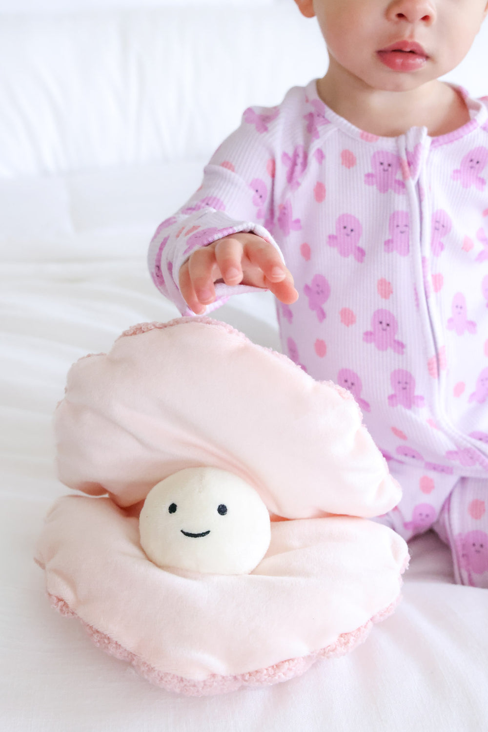 baby in sea snuggles bamboo waffle convertible zip romper reaching for pink clamshell plush toy on white bedding