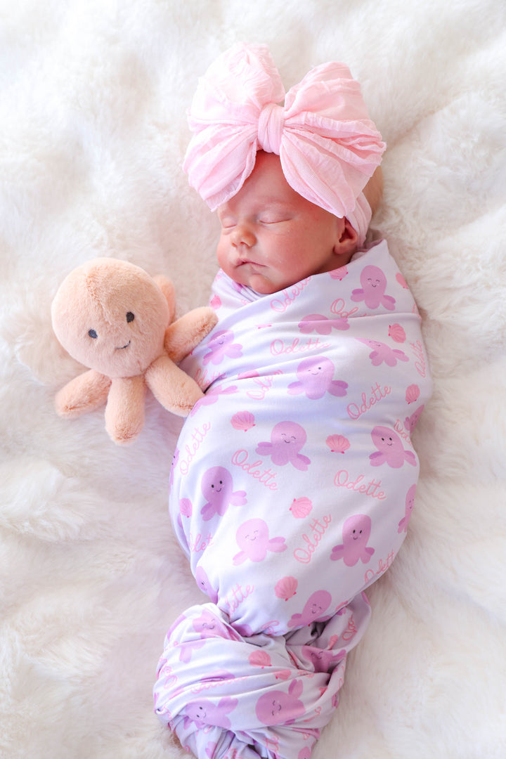 newborn in personalized sea snuggles swaddle blanket with custom name and pink octopus plush toy beside them on white fur
