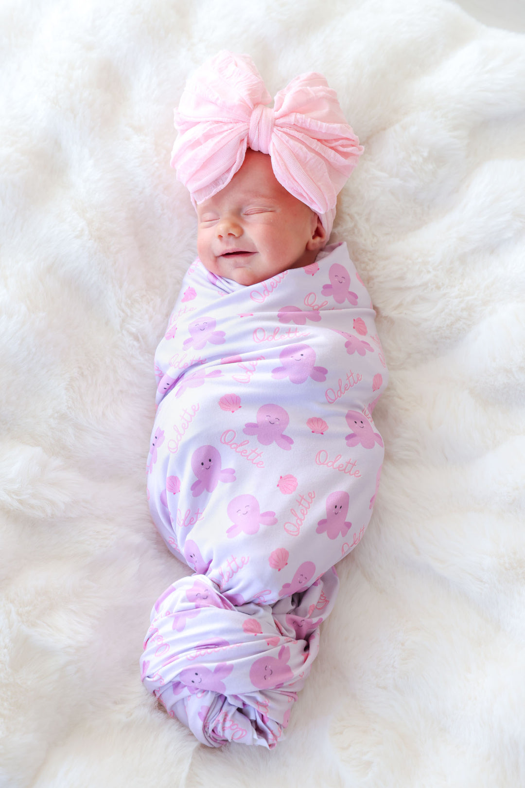 smiling newborn swaddled in personalized sea snuggles swaddle blanket in lavender with octopus and seashell print and custom name — with pink tulle bow headband