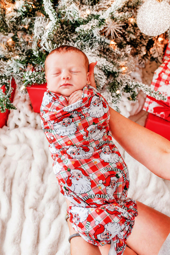 Personalized Cozy Claus Christmas bamboo swaddle blanket featuring a vintage Santa print, wrapped around a sleeping newborn for Christmas newborn photos and personalized baby gifts.