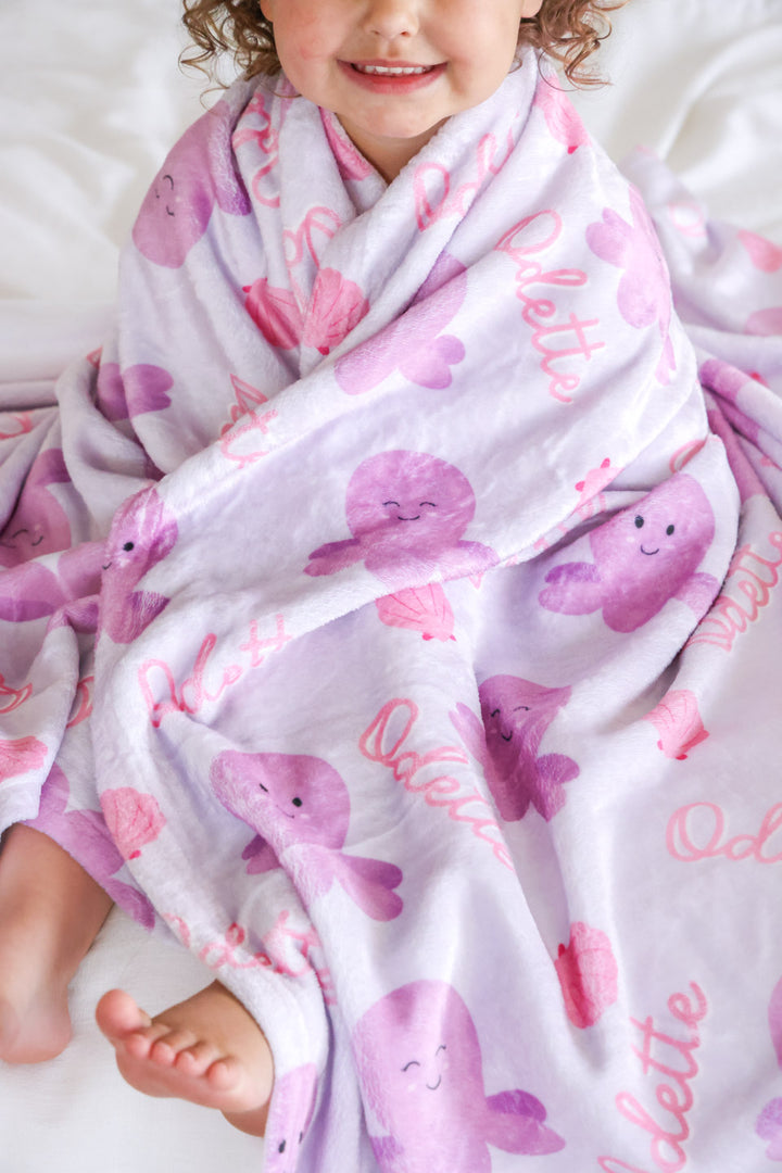 personalized sea snuggles kids blanket folded on white background — showing octopus and seashell print with custom name in pink script