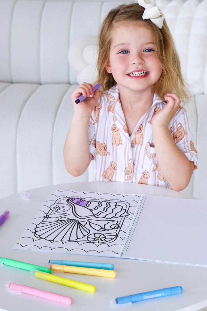 Smiling young girl with a bow in her hair holding a purple marker, seated at a table with a partially colored seashell page from a Caden Lane coloring book and an array of colorful markers