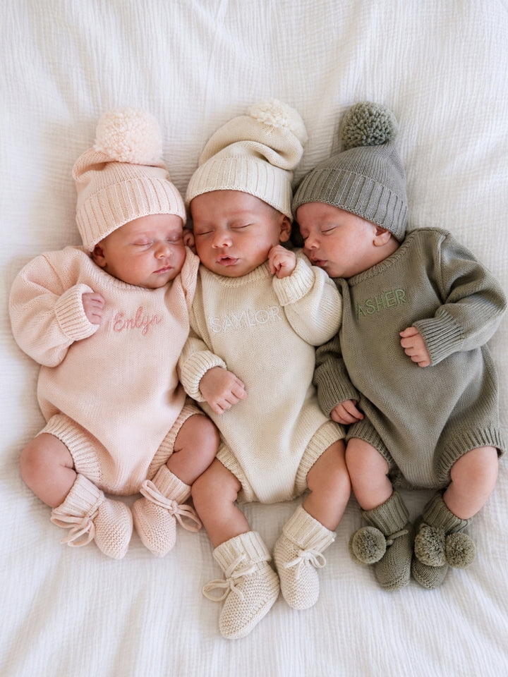 Three newborn babies lying side by side on a white textured blanket, wearing Caden Lane knit rompers, hats, and booties in soft blush pink, cream, and sage green. 