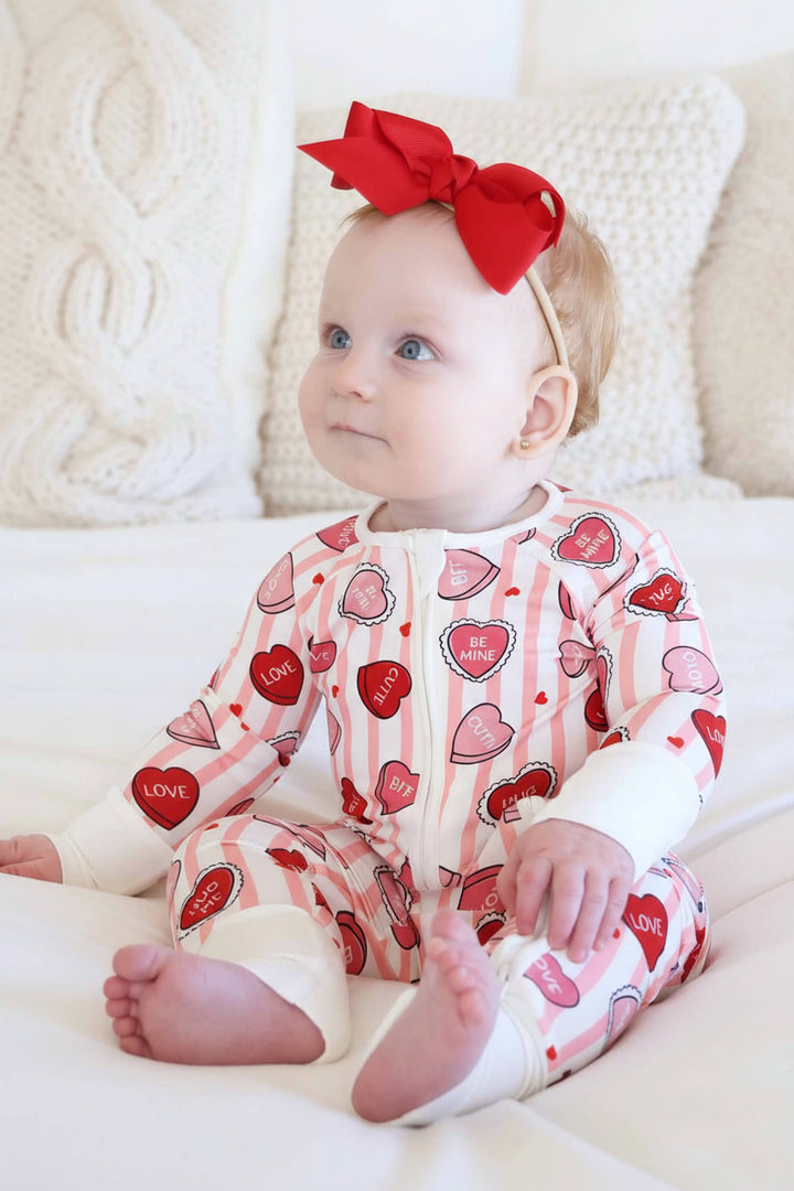 baby girl  sitting on a bed wearing a pink stripe candy heart convertible zip romper pajama 