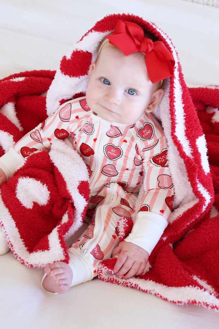 baby girl wearing a pink and white stripe convertible zip romper with pink and red conversation heart candy with a heart plush blanket over her head 