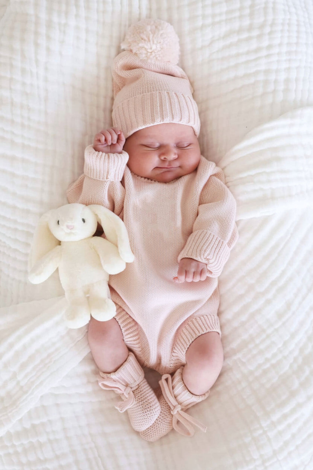Sleeping baby wearing Caden Lane blush pink knit sweater romper with matching pom-pom hat and tie booties, next to a plush bunny