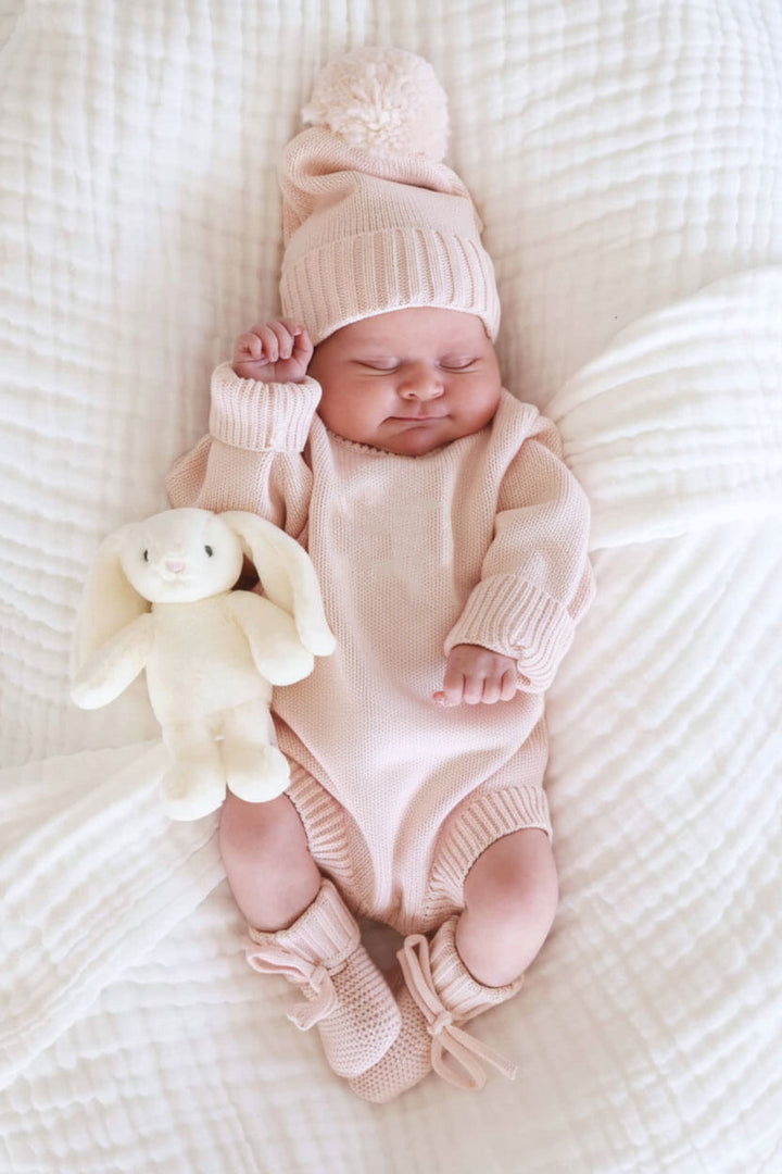 Sleeping baby wearing Caden Lane blush pink knit sweater romper with matching pom-pom hat and tie booties, next to a plush bunny