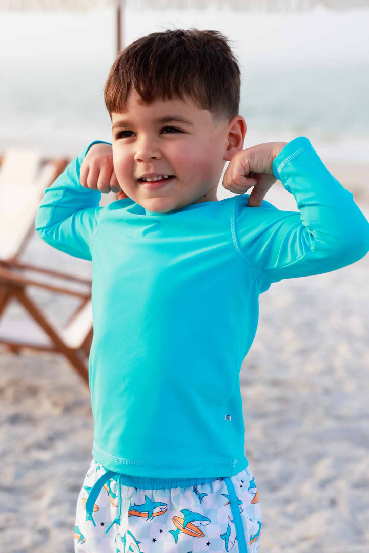 boy flexing arms at the beach in blue long sleeve rash guard shirt 