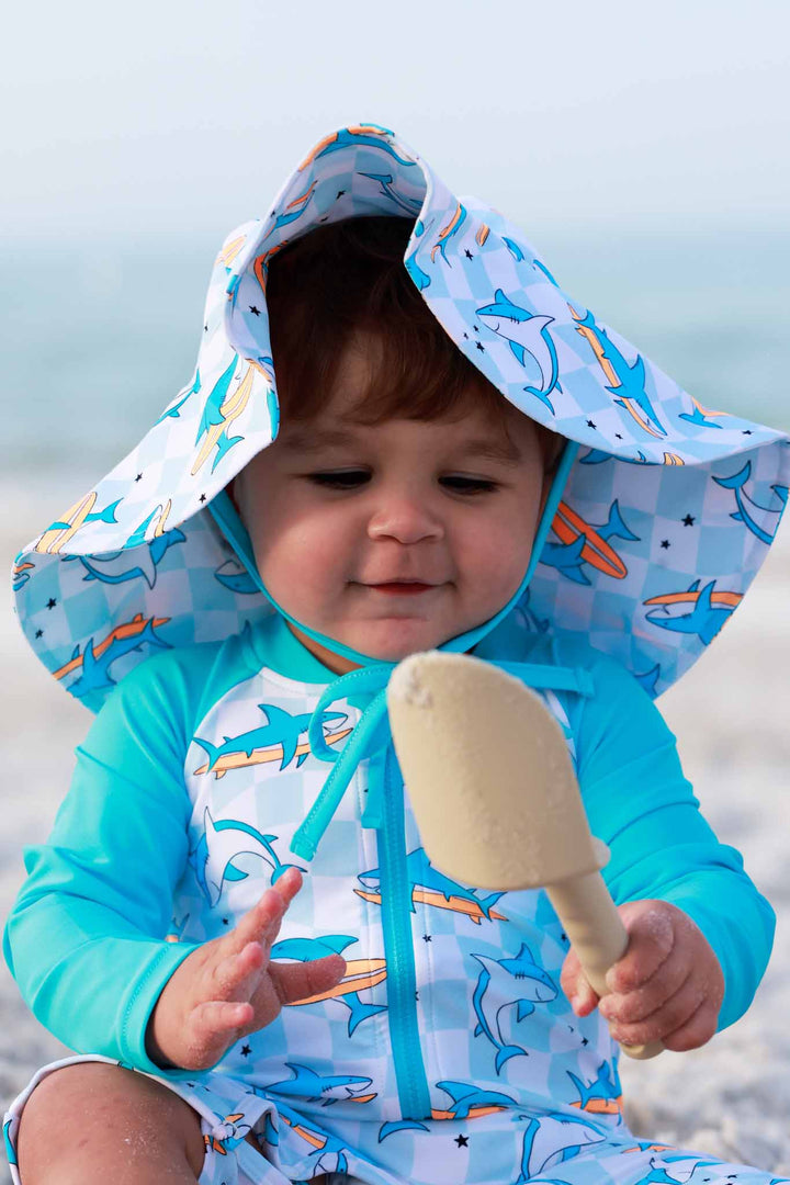 boy sitting on beach in surfing shark rash guard romper with matching sun hat 