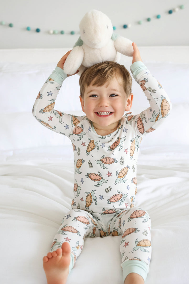 boy sitting on bed in turtle pajama set with  a stuffed turtle on his head 