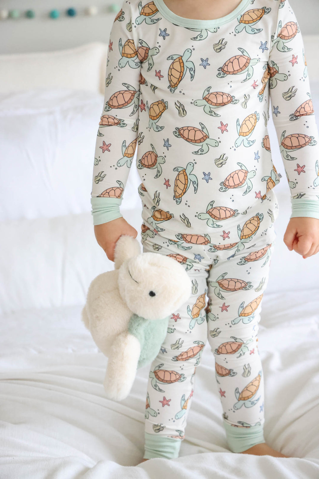kid standing on bed in turtle pajamas holding a large turtle stuffed animal 
