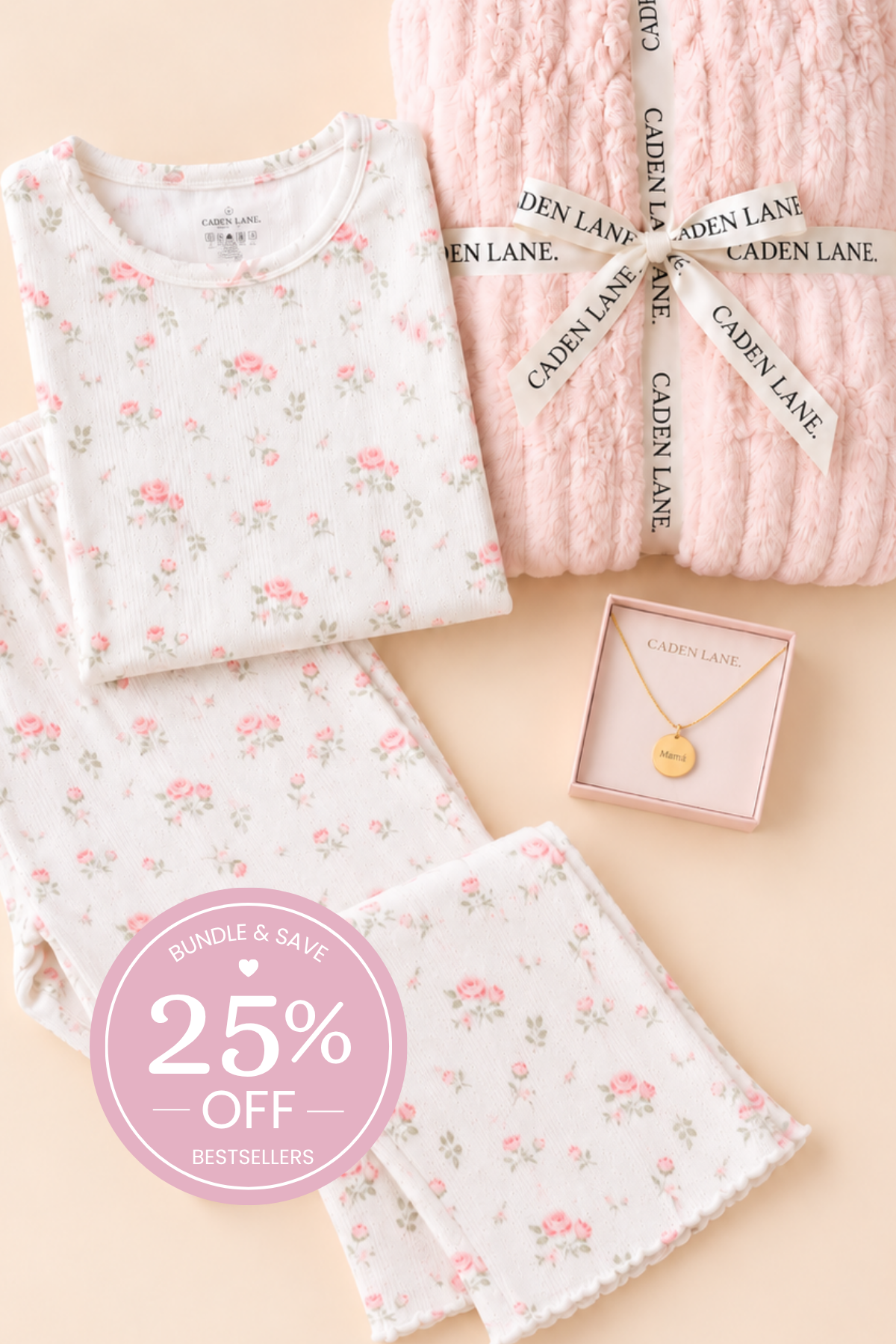 A flat-lay of Caden Lane mom ultimate gift set,  featuring a white long-sleeve top and matching pants with a pink and green floral print, next to a chunky light pink knit blanket tied with a branded ribbon, and a small gold pendant necklace in a pink box. A pink circular badge in the corner advertises a 25% off bundle.